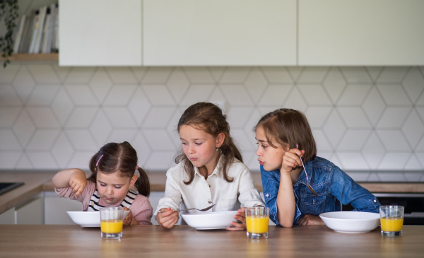 A portrait of three girls sisters indoors at home, eating breakfast.