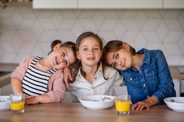 A portrait of three girls sisters indoors at home, looking at camera when eating breakfast.