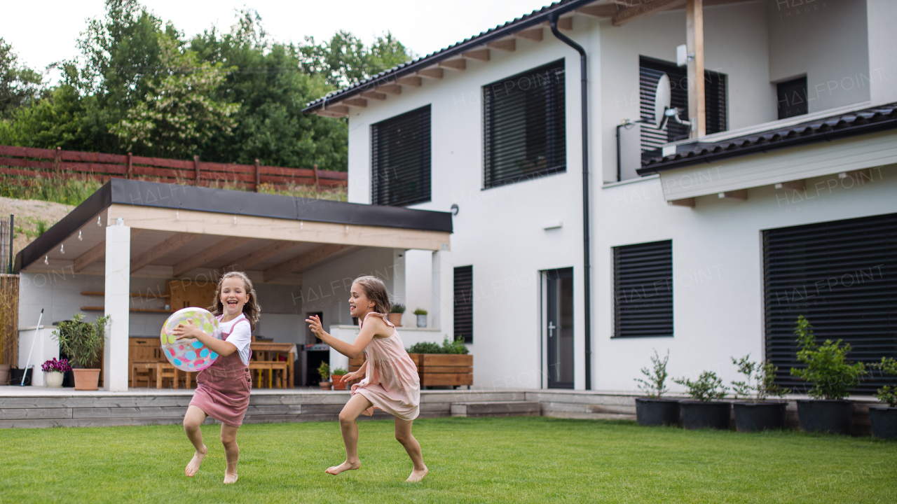 Two happy small girls sisters playing with a ball outdoors in the backyard, running.