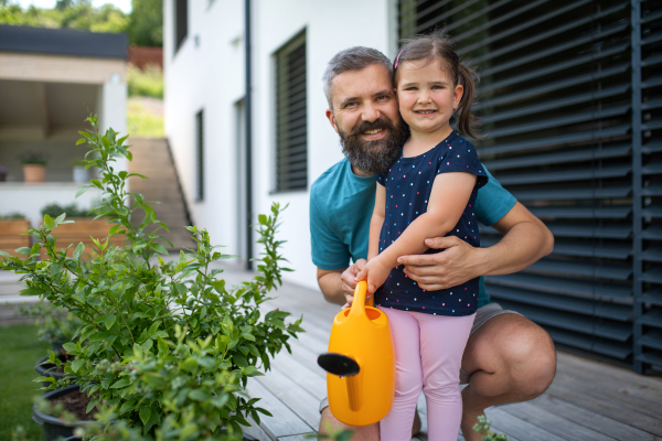 A father and small daughter with watering can outdoors in tha backyard, looking at camera.