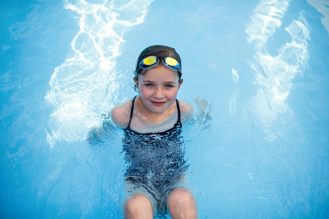 A top view of small girl outdoors in the backyard, sitting in swimming pool and looking at camera.