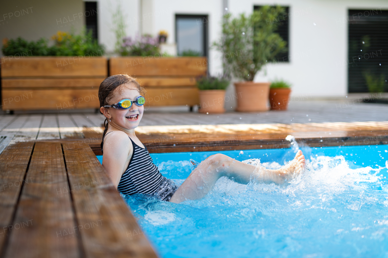 A small girl with goggles outdoors in the backyard, sitting in swimming pool and looking at camera.