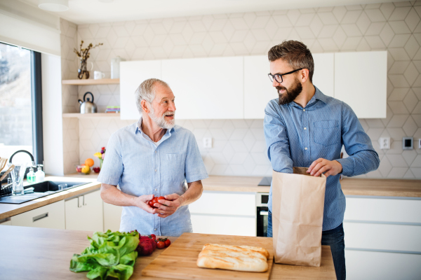 A cheerful adult hipster son and senior father indoors in kitchen at home, unpacking shopping.