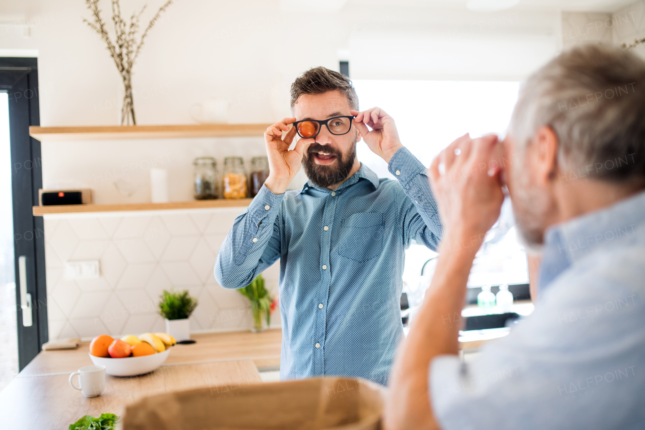 An adult hipster son and senior father indoors in kitchen at home, having fun.