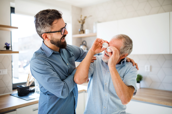 An adult hipster son and senior father with laptop indoors in kitchen at home, having fun.