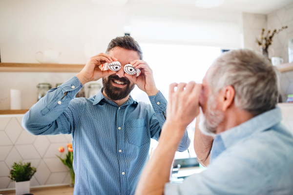 A portrait of adult hipster son and senior father standing indoors at home, having fun.