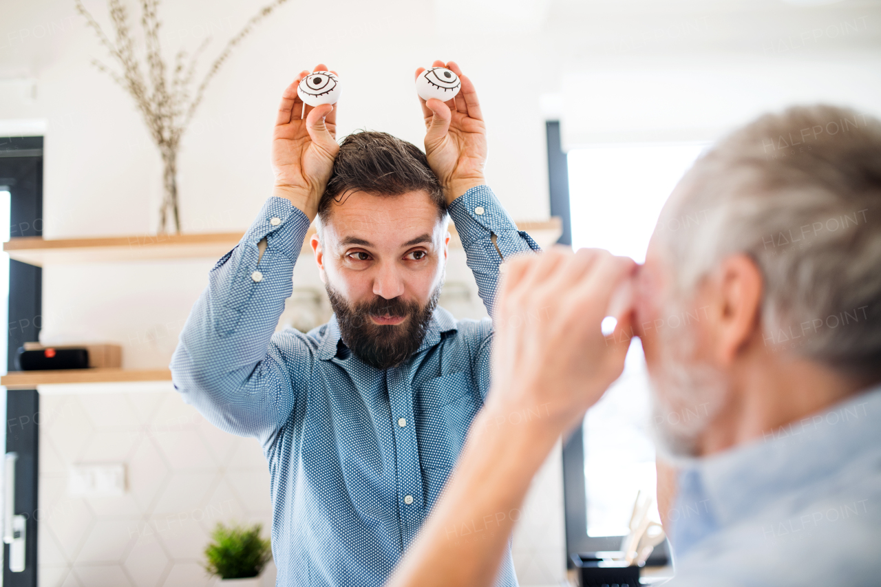 An adult hipster son and senior father indoors in kitchen at home, having fun.