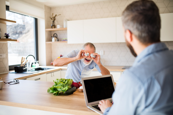 An adult hipster son and senior father with laptop indoors in kitchen at home, having fun.