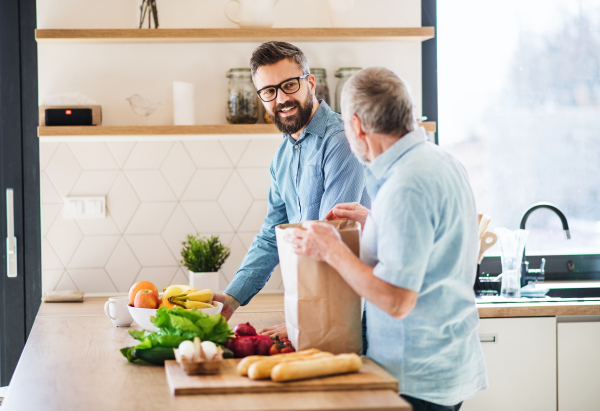 A cheerful adult hipster son and senior father indoors in kitchen at home, unpacking shopping.