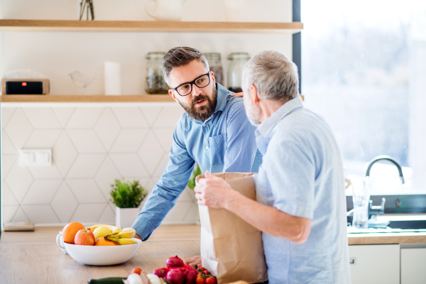 A cheerful adult hipster son and senior father indoors in kitchen at home, unpacking shopping.