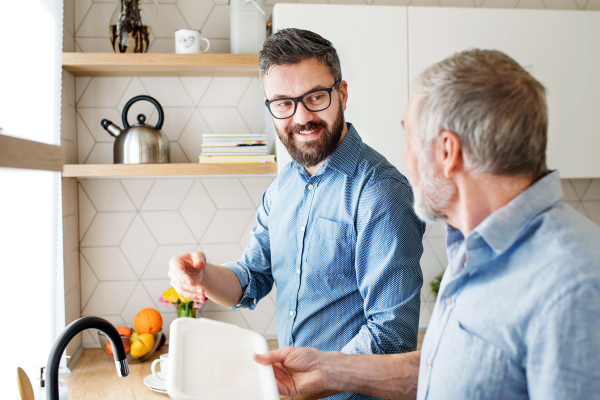 An adult hipster son and senior father indoors in kitchen at home, washing dishes.