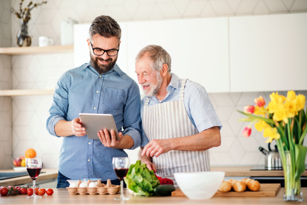 A portrait of adult hipster son and senior father indoors in kitchen at home, using tablet.