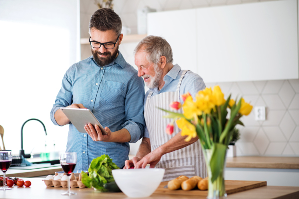 A portrait of adult hipster son and senior father cooking indoors in kitchen at home, using tablet.