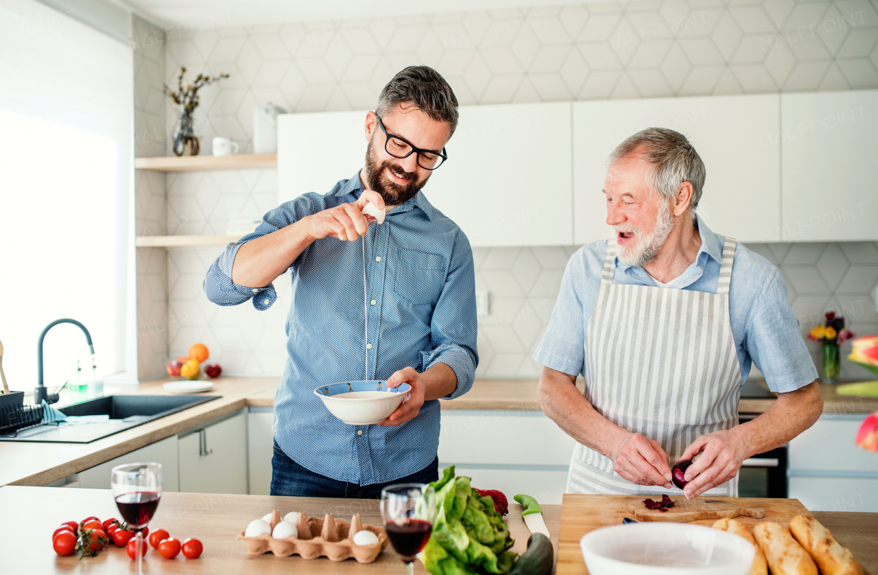 An adult hipster son and senior father indoors in kitchen at home, cooking.