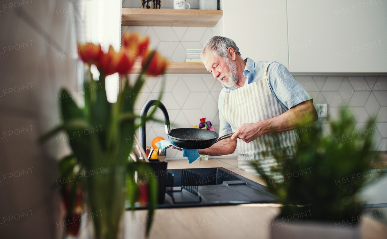 A senior man indoors in kitchen at home, washing up a pan after cooking.