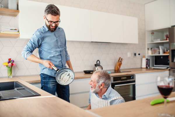 An adult hipster son and senior father indoors in kitchen at home, cooking.