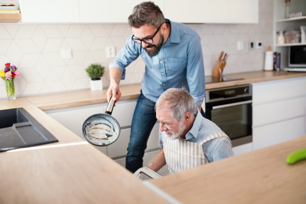 An adult hipster son and senior father indoors in kitchen at home, cooking.