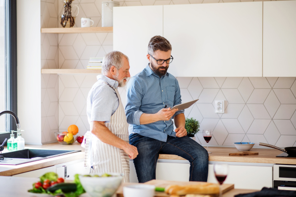 A portrait of adult hipster son and senior father indoors in kitchen at home, using tablet.