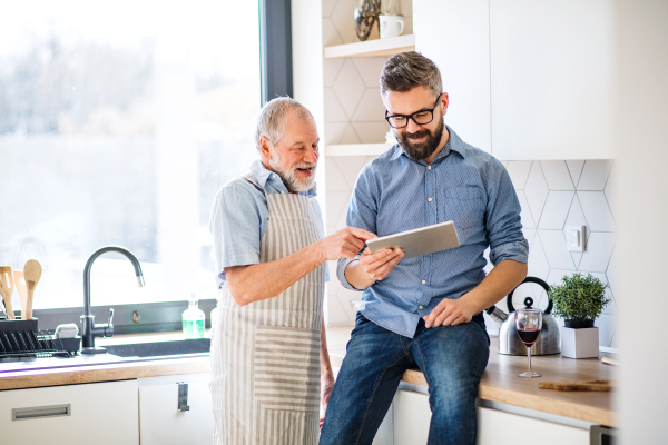 A portrait of adult hipster son and senior father indoors in kitchen at home, using tablet.