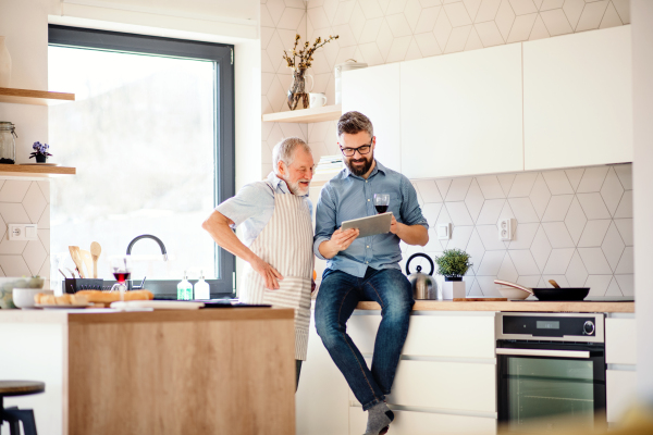 A portrait of adult hipster son and senior father indoors in kitchen at home, using tablet.