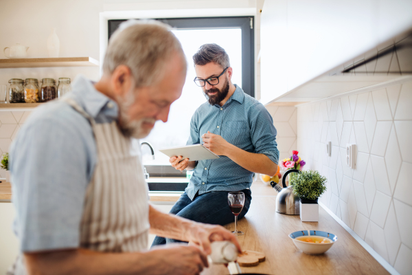 A portrait of adult hipster son and senior father indoors in kitchen at home, using tablet.