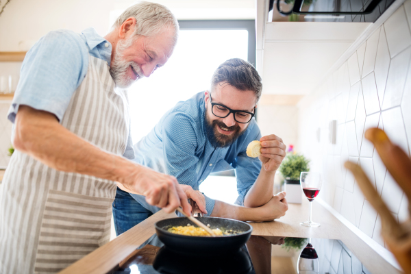 An adult hipster son and senior father indoors in kitchen at home, cooking.