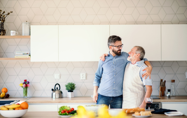 A portrait of adult hipster son and senior father indoors in kitchen at home, talking.