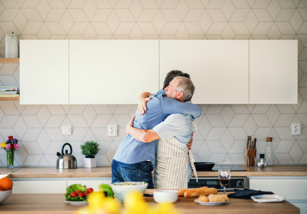 An adult hipster son and senior father indoors in kitchen at home, hugging when cooking.