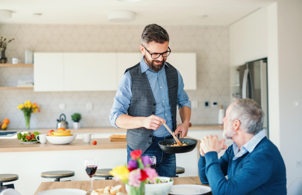 An adult hipster son and senior father indoors in kitchen at home, cooking.