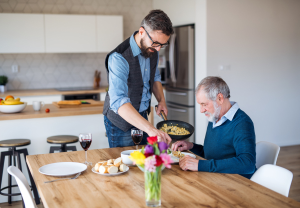 Adult hipster son and senior father sitting at the table indoors at home, having lunch.