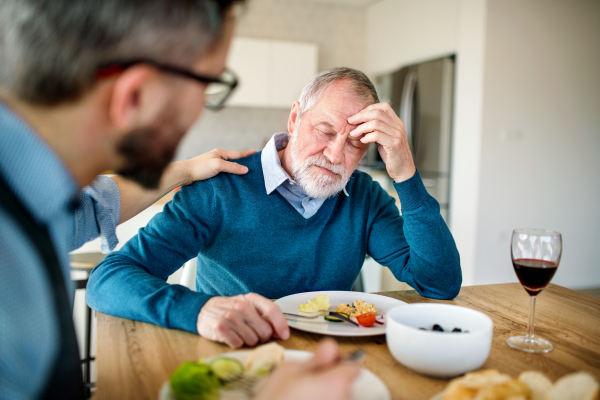 An adult hipster son comforting frustrated senior father indoors at home, eating light lunch.