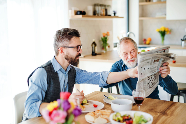 An adult hipster son and senior father sitting at the table indoors at home, eating light lunch and reading newspapers.