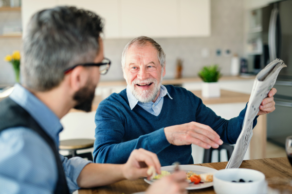 An adult hipster son and senior father sitting at the table indoors at home, eating light lunch.