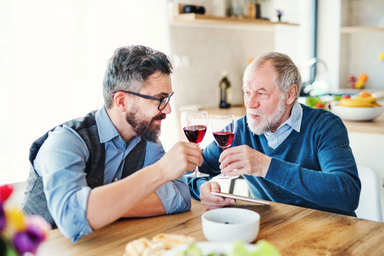 An adult hipster son and senior father sitting at the table indoors at home, eating light lunch.