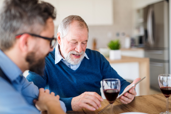 Adult hipster son and senior father sitting at the table indoors at home, using tablet.