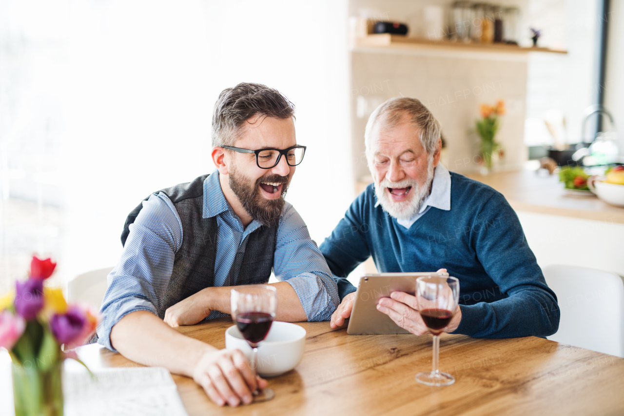 Adult hipster son and senior father with wine and tablet sitting at the table indoors at home.