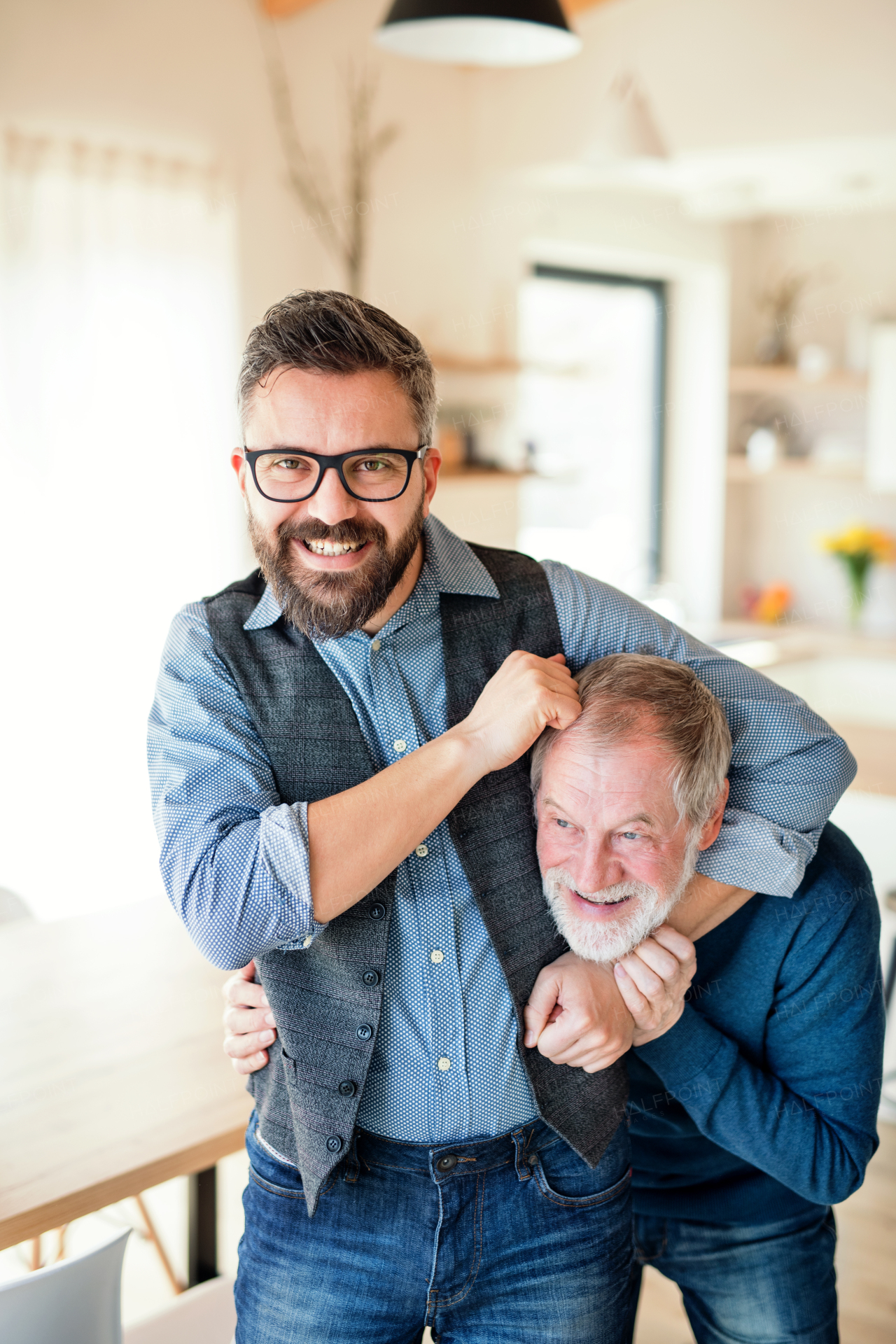 An adult hipster son with glasses and senior father indoors at home, having fun.