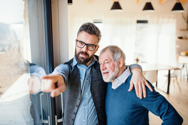 A portrait of adult hipster son and senior father standing indoors at home, talking.