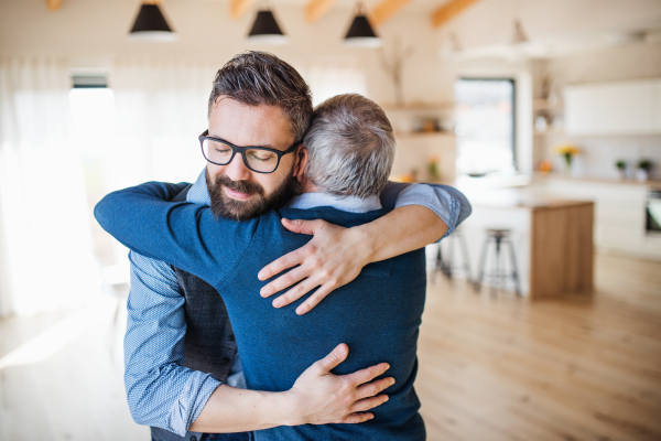 An adult son and senior father standing indoors at home, hugging.