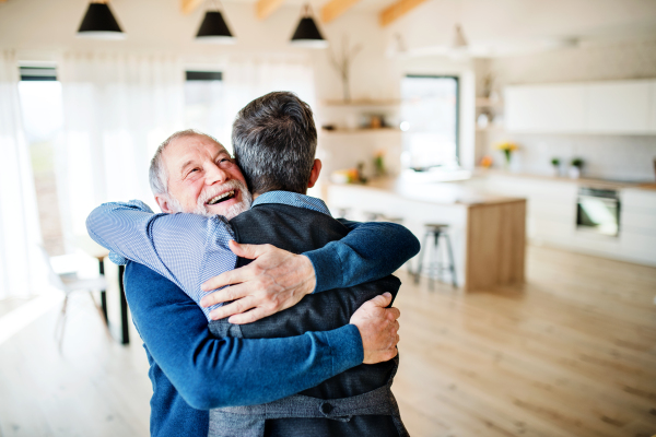 An adult hipster son with glasses and senior father indoors at home, hugging.