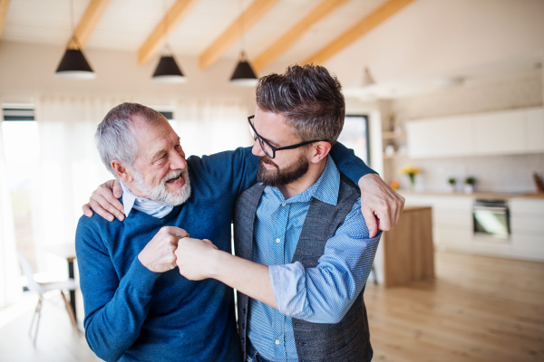 An adult son and senior father standing arm in arm indoors at home, making fist bump.