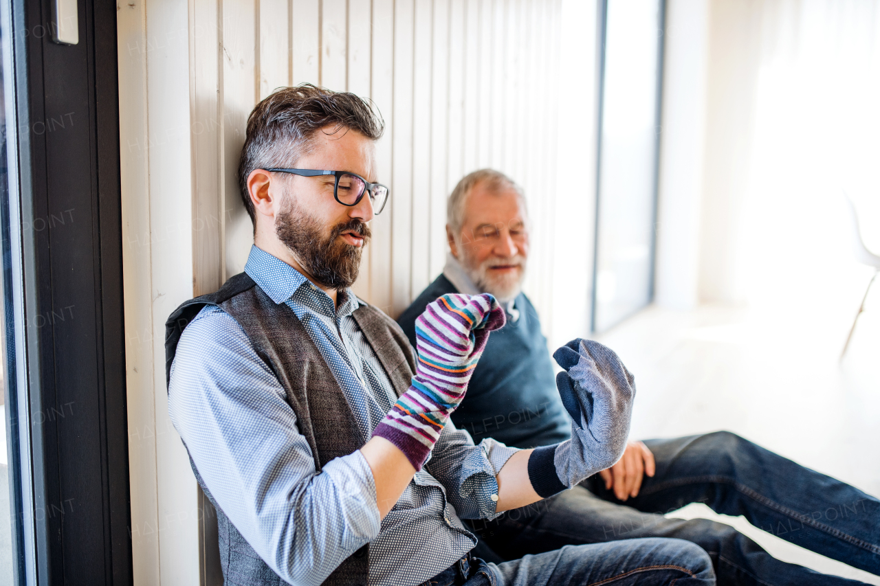 A portrait of adult hipster son and senior father sitting on floor indoors at home, having fun.