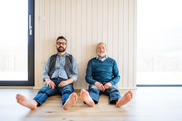 A portrait of adult hipster son and senior father sitting on floor indoors at home, white wooden wall in the background.