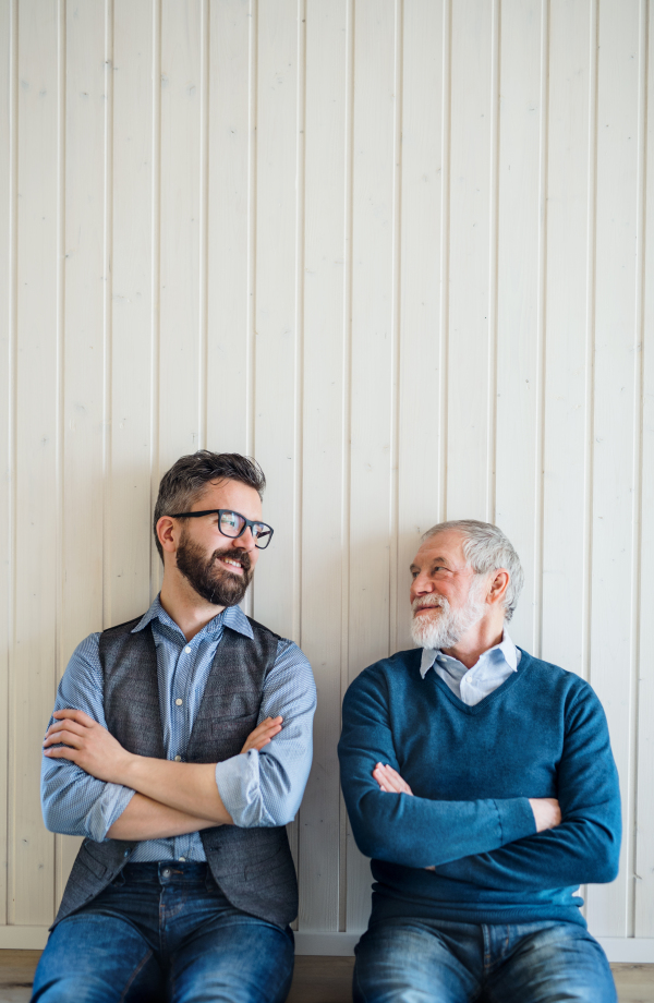 A portrait of adult hipster son and senior father sitting on floor indoors at home, white wooden wall in the background.