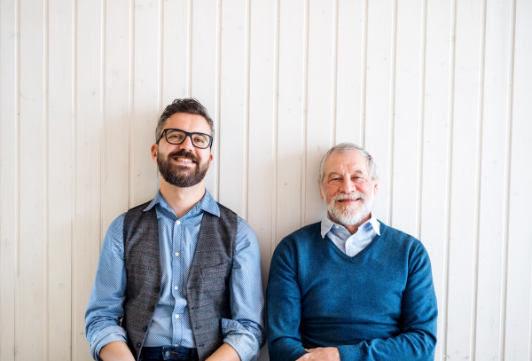 A portrait of adult hipster son and senior father sitting on floor indoors at home, white wooden wall in the background.