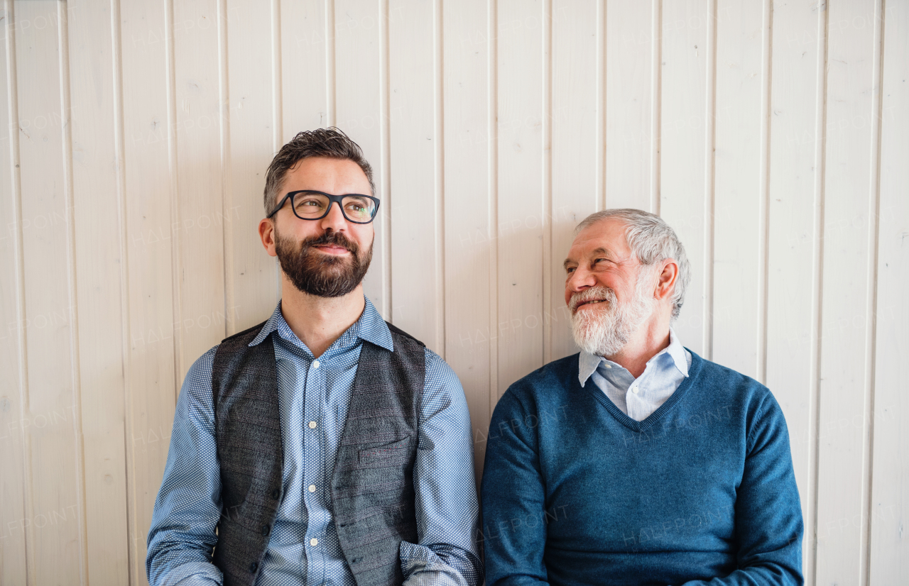 A portrait of adult hipster son and senior father sitting on floor indoors at home, white wooden wall in the background.