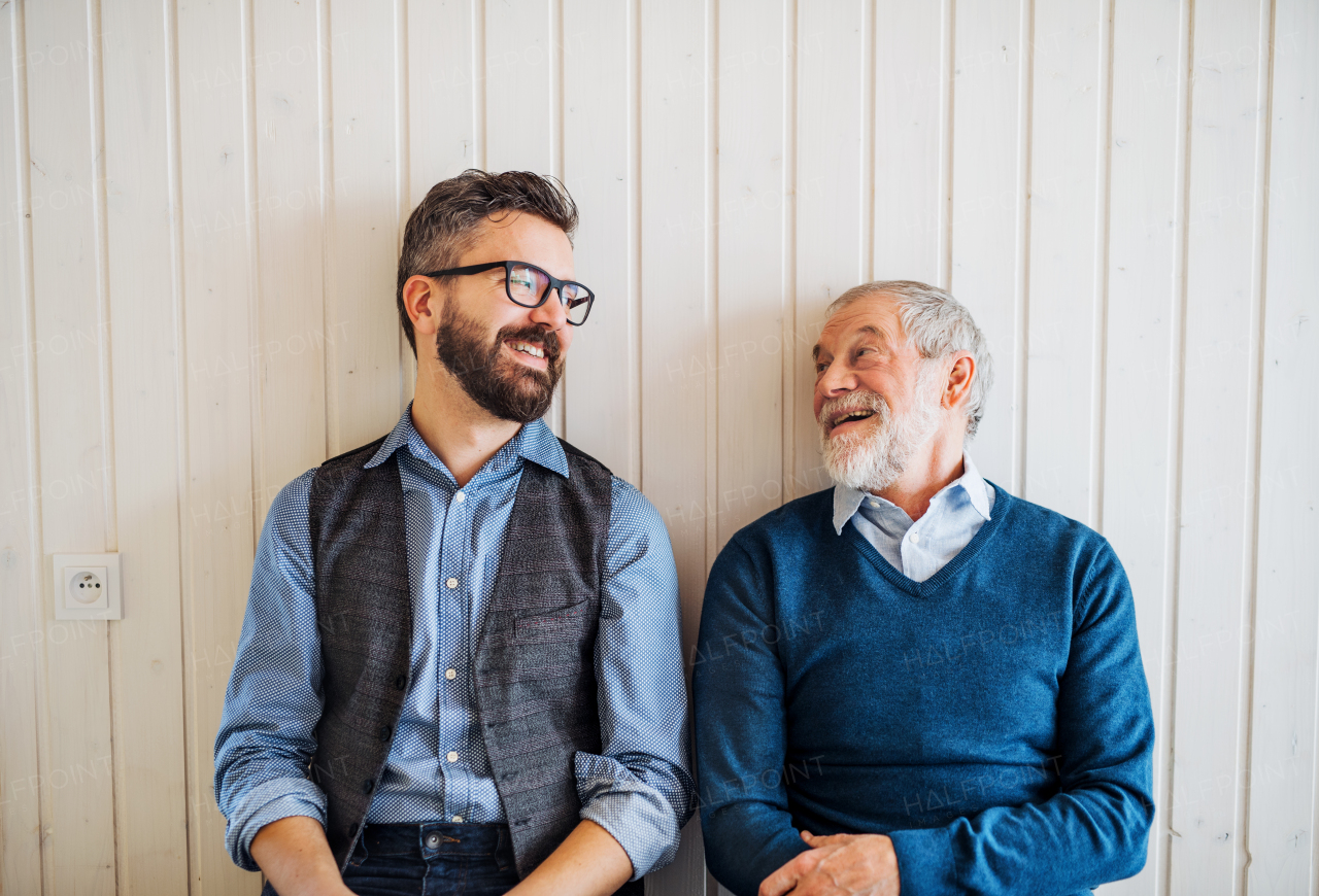 A portrait of adult hipster son and senior father sitting on floor indoors at home, white wooden wall in the background.