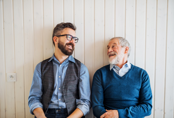 A portrait of adult hipster son and senior father sitting on floor indoors at home, white wooden wall in the background.