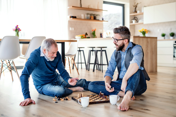 A cheerful adult hipster son and senior father sitting on floor indoors at home, playing chess.