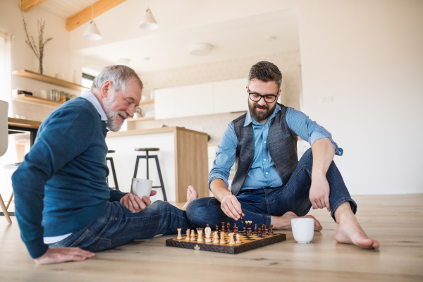 A cheerful adult hipster son and senior father sitting on floor indoors at home, playing chess.